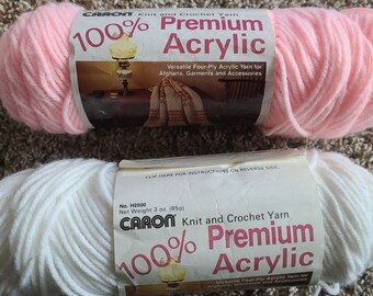 May include: Two skeins of Caron knit and crochet yarn, one light pink and one white. Each label reads 100% Premium Acrylic. The white skein label specifies a net weight of 3 oz. (85g). Both labels feature a small image of a lamp and a blanket.