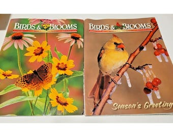May include: Two Birds & Blooms magazines side by side. The left cover features a butterfly on yellow flowers and pink coneflowers. The right cover shows a yellow cardinal on a branch with red berries and the text Season's Greeting.