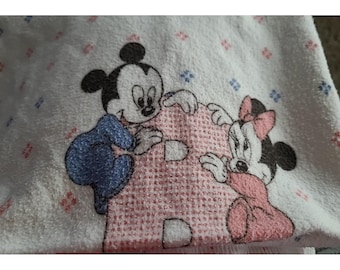May include: A white fabric towel featuring an embroidered design of Mickey Mouse in a blue outfit and Minnie Mouse in a pink outfit, both positioned around a large, textured pink letter B. The background has small red and blue floral accents.