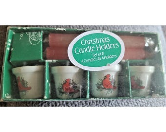 May include: A boxed set of Christmas Candle Holders containing four white ceramic candle holders decorated with red cardinal birds and green pine branches, along with four red taper candles. The green packaging features the text Christmas Candle Holders.