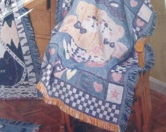 May include: A woven throw blanket featuring a blue and cream color palette with a central illustration of two teddy bears wearing bow ties. The design includes the word BABY, checkered patterns, hearts, and stars, finished with a fringed edge.