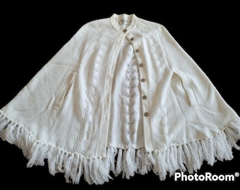 Könnte beinhalten: Cremefarbene Strick-Cape mit rundem Kragen und Knopfverschluss. Die Cape hat blattförmige Aussparungen, Taschen und eine Fransenborte. Ein Kleidungsstück im Vintage-Stil.
