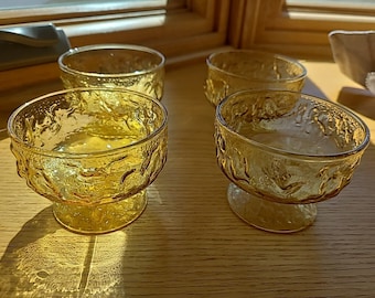 Könnte beinhalten: Vier bernsteinfarbene Dessertschalen aus Glas mit strukturierter Oberfläche. Jede Schale hat einen Fuß und ein strukturiertes Design am Rand. Die Schalen stehen auf einer Holzoberfläche, mit Schatten durch Sonnenlicht.