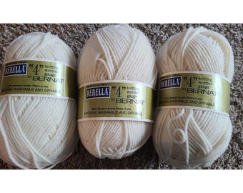 May include: Three skeins of off-white Berella 4 knitting worsted gauge yarn by Bernat. Each skein features a gold label with black text stating 100% blended acrylic fibers, 4 ply, and machine washable and dryable. The yarn is a solid cream color.
