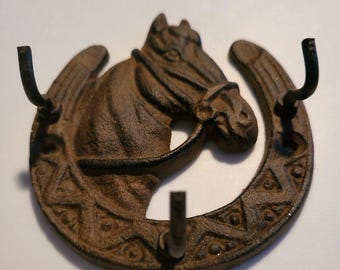May include: A dark brown cast iron wall hook featuring a horse head centered within a horseshoe frame. The horseshoe is decorated with a repeating triangular and dot pattern and includes three upward-curving hooks for hanging items.