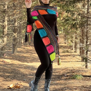 May include: A woman wearing a black turtleneck shirt and black leggings with a long, colorful crochet scarf. The scarf has a geometric pattern with squares of pink, orange, blue, green, and yellow. The woman is wearing black boots and sunglasses.