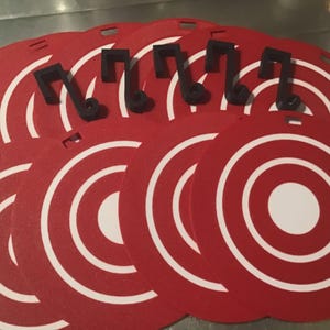 May include: A set of circular red and white target-patterned mats, each featuring a small rectangular cutout at the top edge. Accompanying the mats are several black S-shaped metal hooks designed for hanging the circular items.