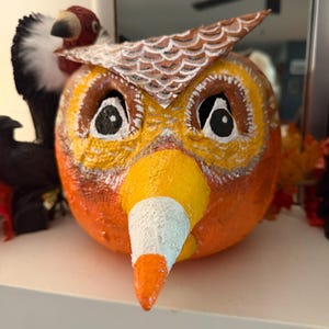 May include: A painted pumpkin decorated to resemble an owl with a large, pointed candy corn-shaped beak. The pumpkin features orange, yellow, and white colors, with detailed painted feathers on the top and large, expressive eyes with black pupils.