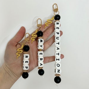 May include: Three keychains with gold-colored snake charms and gold-colored clasps. The keychains have black and white beads spelling out "REP", "MY ERA", and "REPUTATION". The keychains are of varying lengths.