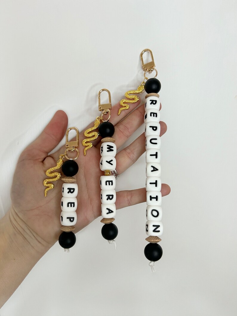 May include: Three keychains with gold-colored snake charms and gold-colored clasps. The keychains have black and white beads spelling out "REP", "MY ERA", and "REPUTATION". The keychains are of varying lengths.