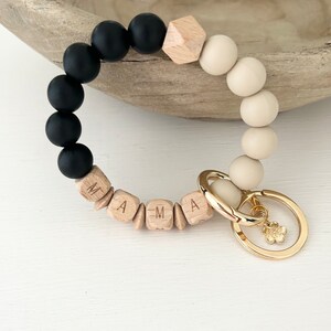 May include: A wristlet keychain with black, beige, and wooden beads. The word "MAMA" is spelled out in wooden letter beads. A gold-colored clasp and flower charm complete the design.