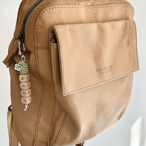 K&ouml;nnte beinhalten: Ein hellbrauner Lederrucksack mit einer Vordertasche und einem silberfarbenen Rei&szlig;verschluss. Ein Schl&uuml;sselanh&auml;nger mit dem Wort "MAMA" auf Holzperlen h&auml;ngt am Rei&szlig;verschluss. Der Schl&uuml;sselanh&auml;nger hat auch einen gr&uuml;nen und silbernen Blumenanh&auml;nger.