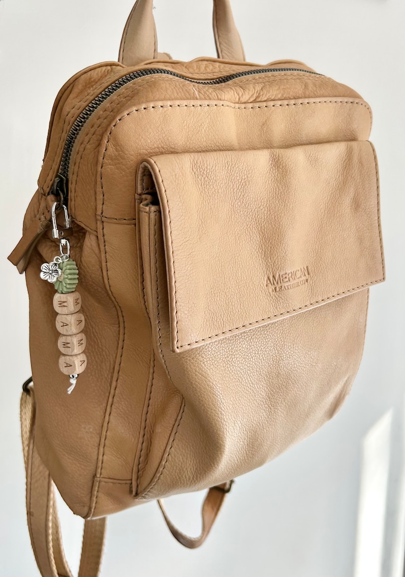 K&ouml;nnte beinhalten: Ein hellbrauner Lederrucksack mit einer Vordertasche und einem silberfarbenen Rei&szlig;verschluss. Ein Schl&uuml;sselanh&auml;nger mit dem Wort "MAMA" auf Holzperlen h&auml;ngt am Rei&szlig;verschluss. Der Schl&uuml;sselanh&auml;nger hat auch einen gr&uuml;nen und silbernen Blumenanh&auml;nger.