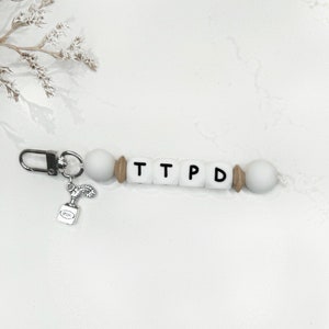 May include: A white beaded keychain with the letters "TTPD" spelled out in black. The keychain has a silver clasp and a small charm of an ink bottle. The beads are white, with wooden and white accents.