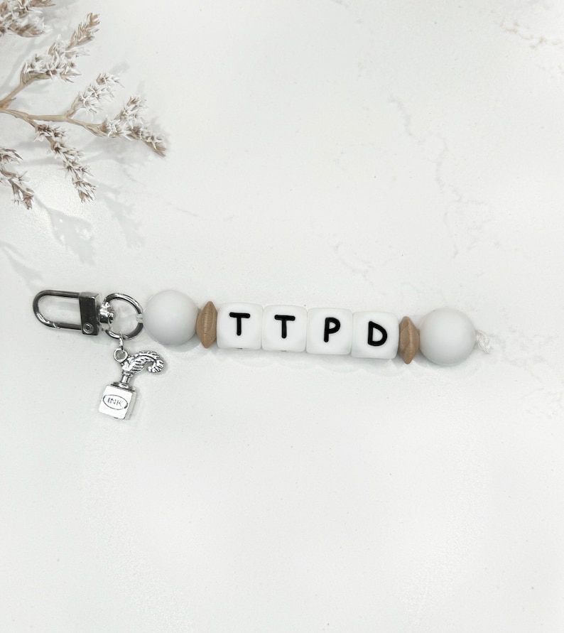 May include: A white beaded keychain with the letters "TTPD" spelled out in black. The keychain has a silver clasp and a small charm of an ink bottle. The beads are white, with wooden and white accents.