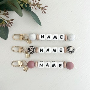 May include: Three personalized keychains with the name "NAME" spelled out in white letter beads. Each keychain has a gold clasp and decorative beads in speckled, leopard print, and mauve colors. A floral charm is attached.