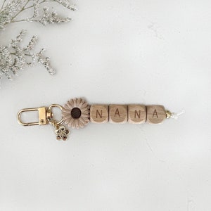 May include: A wooden keychain with the name "NANA" spelled out in individual letter blocks. The keychain features a gold-colored clasp, a small flower charm, and a beige sunflower accent. The background is white.
