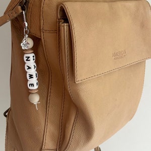 May include: A tan leather backpack with a silver zipper and a small pocket. Attached is a beaded keychain spelling "NAME" in black letters on white beads, with wooden and pearl accents, and a silver flower charm.