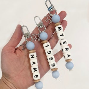 May include: Three personalized keychains with white lettered beads spelling out names and numbers. Each keychain has a silver clasp, a light blue bead, and a wooden bead. One keychain says "NAME" and another says "1989".