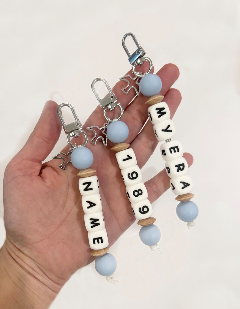 May include: Three personalized keychains with white lettered beads spelling out names and numbers. Each keychain has a silver clasp, a light blue bead, and a wooden bead. One keychain says "NAME" and another says "1989".