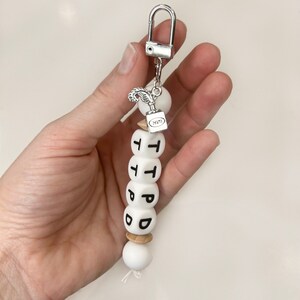 May include: A white beaded keychain with black lettered beads spelling out "TPPD". The keychain has a silver clasp and a small charm with a feather and inkwell design. The beads are strung together with a white cord.