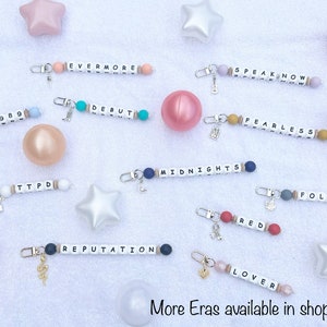 May include: Keychains with white letter beads spelling out album titles like "Evermore," "Speak Now," and "Debut." Each keychain has a charm and colorful beads. The text "More Eras available in shop" is at the bottom.