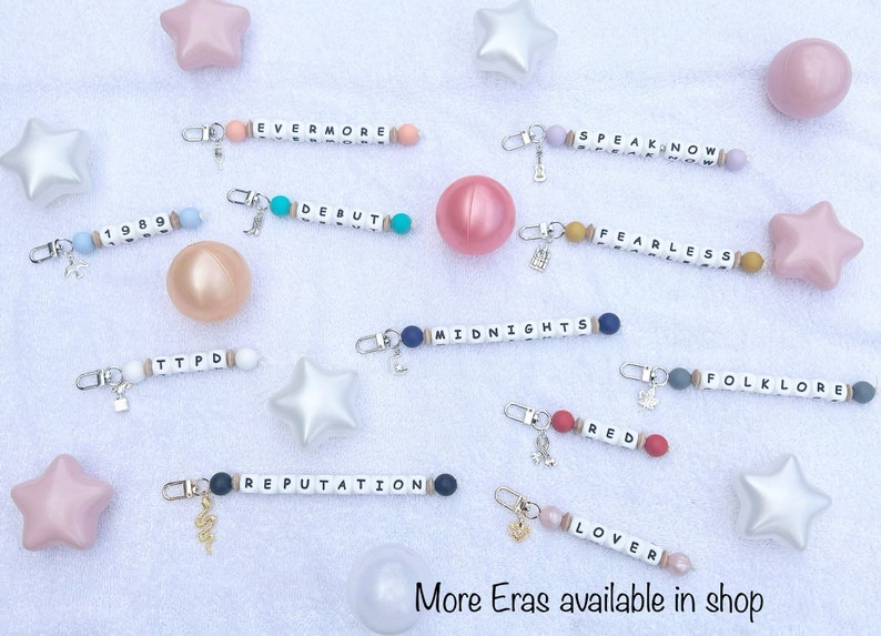 May include: Keychains with white letter beads spelling out album titles like "Evermore," "Speak Now," and "Debut." Each keychain has a charm and colorful beads. The text "More Eras available in shop" is at the bottom.