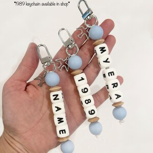 May include: Three personalized keychains with white lettered beads spelling out names and numbers. Each keychain has a silver clasp, a light blue bead, and a wooden bead. The text "Examples of different wording" is at the top.