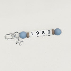 May include: A beaded keychain with the year "1989" spelled out in white letter beads. The keychain features blue and wooden beads, a silver bird charm, and a silver clasp.