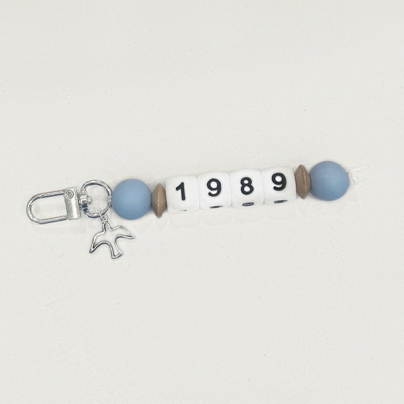 May include: A beaded keychain with the year "1989" spelled out in white letter beads. The keychain features blue and wooden beads, a silver bird charm, and a silver clasp.