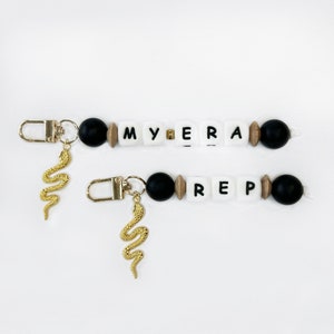 May include: Two beaded keychains with gold-colored snake charms. One keychain spells "MYERA" with white letter beads, black beads, and wooden beads. The other keychain spells "REP" with similar beads. Both have gold-colored clasps.