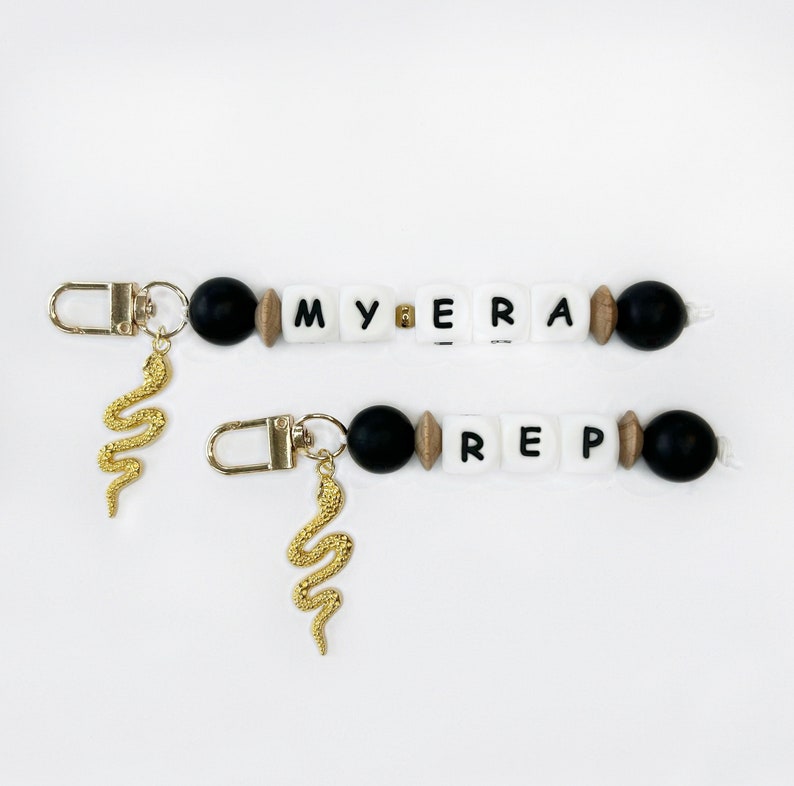 May include: Two beaded keychains with gold-colored snake charms. One keychain spells "MYERA" with white letter beads, black beads, and wooden beads. The other keychain spells "REP" with similar beads. Both have gold-colored clasps.