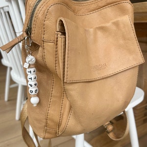 May include: A tan leather backpack with a front pocket and a zipper closure. The backpack has a small keychain with the letters T P D D on it. The backpack is sitting on a white chair.