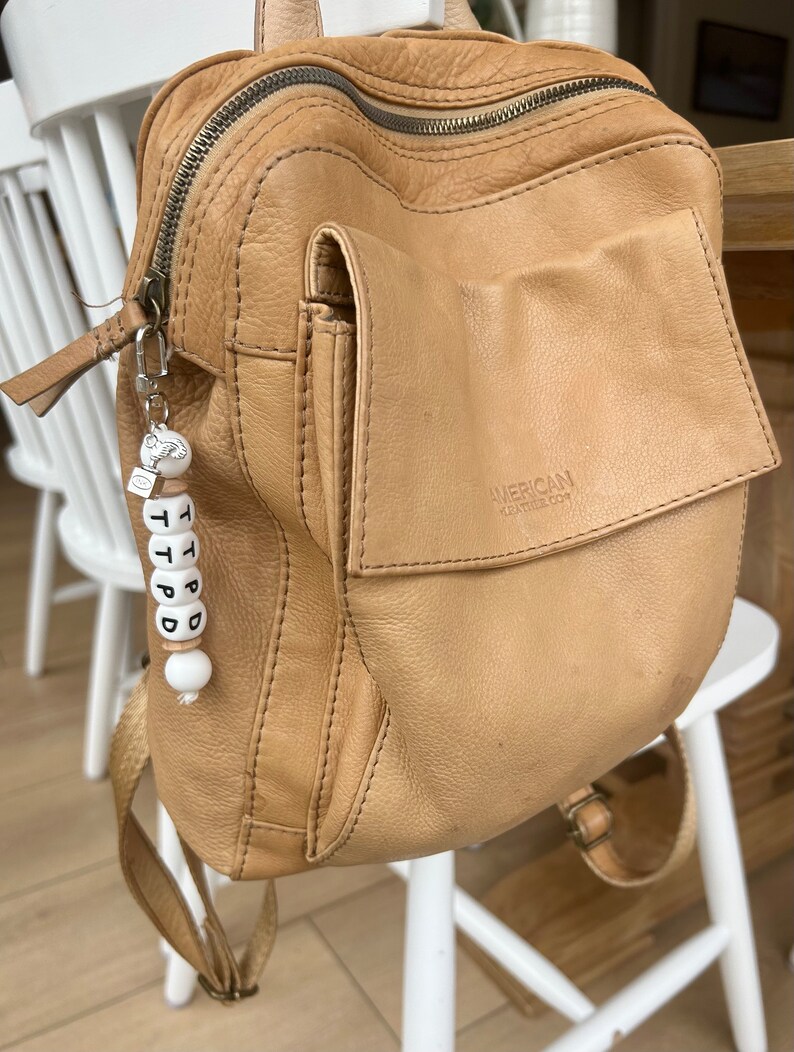 May include: A tan leather backpack with a front pocket and a zipper closure. The backpack has a small keychain with the letters T P D D on it. The backpack is sitting on a white chair.