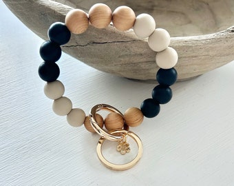 May include: A wooden bead keychain bracelet in a circular design. The bracelet features a mix of natural wood, black, and cream-colored beads. A gold-colored clasp and key ring with a small flower charm are attached.