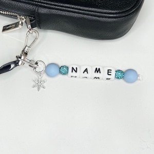 May include: A black leather purse with a silver clasp and a custom beaded keychain. The keychain spells "NAME" with white letter beads, light blue beads, and a silver snowflake charm. The beads are strung on a white cord.