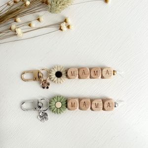 K&ouml;nnte beinhalten: Zwei Schl&uuml;sselanh&auml;nger mit dem Wort "MAMA" aus Holzbuchstaben. Einer hat einen goldenen Verschluss und eine wei&szlig;e G&auml;nsebl&uuml;mchenbl&uuml;te, der andere einen silbernen Verschluss und eine gr&uuml;ne G&auml;nsebl&uuml;mchenbl&uuml;te. Beide haben Blumenanh&auml;nger.