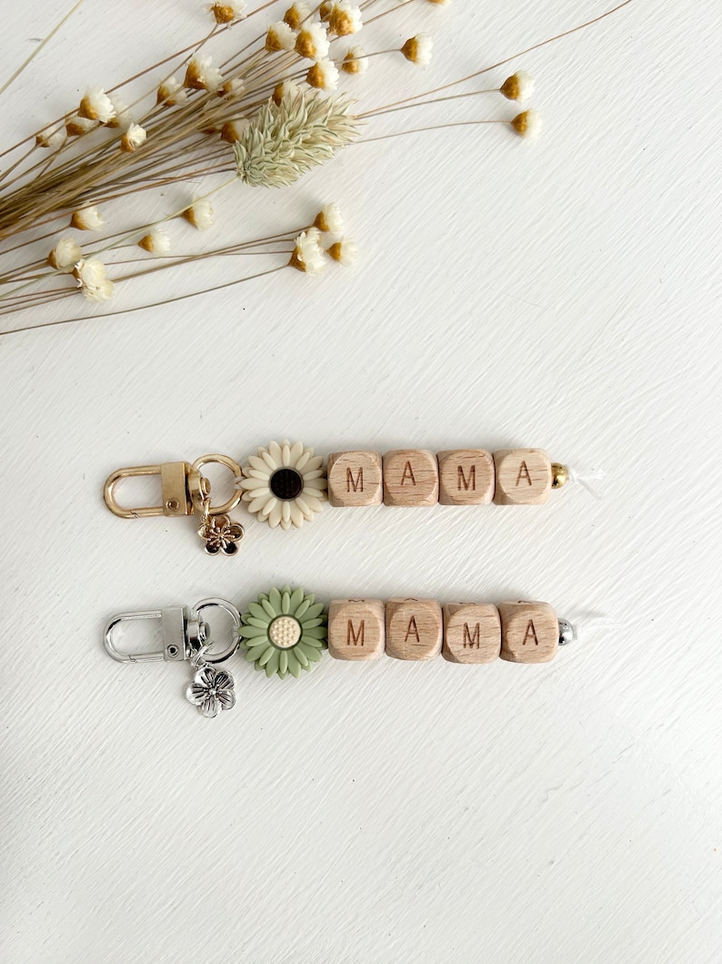 K&ouml;nnte beinhalten: Zwei Schl&uuml;sselanh&auml;nger mit dem Wort "MAMA" aus Holzbuchstaben. Einer hat einen goldenen Verschluss und eine wei&szlig;e G&auml;nsebl&uuml;mchenbl&uuml;te, der andere einen silbernen Verschluss und eine gr&uuml;ne G&auml;nsebl&uuml;mchenbl&uuml;te. Beide haben Blumenanh&auml;nger.