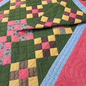 May include: A handmade quilt with a patchwork design. The quilt features squares of green, pink, yellow, and brown fabric. The quilt is bordered with a blue floral pattern and a red background.