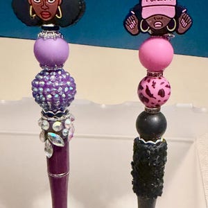 Puede incluir: Dos bolígrafos decorativos con adornos negros estilo afro que llevan gorras con las palabras "Queens" y "Periodt". Los bolígrafos tienen cuentas moradas, rosas y negras, y el bolígrafo morado tiene detalles de pedrería. Los barriles de los bolígrafos son morados y negros.
