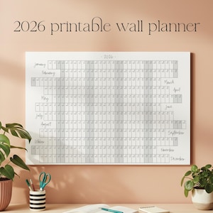 Peut inclure: Un planificateur mural imprimable blanc pour 2026 avec une grille affichant les mois et les dates. Le texte "2026 printable wall planner" est en haut. Le planificateur est affiché sur un mur rose clair, avec des plantes et des fournitures de bureau.