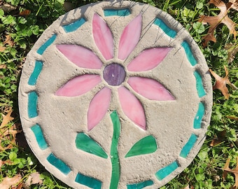 May include: A circular concrete stepping stone featuring a mosaic design of a flower. The flower has light pink stained glass petals surrounding a dark purple center. A green glass stem and leaves are below, with teal glass pieces bordering the edge.