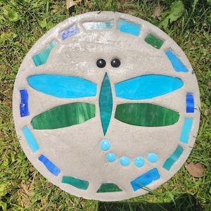 May include: A round, concrete stepping stone with a mosaic dragonfly design. The dragonfly has turquoise and green wings, with a turquoise body and head. The border is decorated with turquoise and green mosaic tiles. The stepping stone is set on green grass.