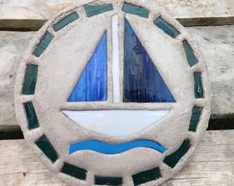 May include: A circular concrete stepping stone featuring a mosaic design of a sailboat. The boat has a white hull, a white mast, and two blue glass sails. A wavy blue glass line represents water below the boat, and dark green glass tiles border the edge.