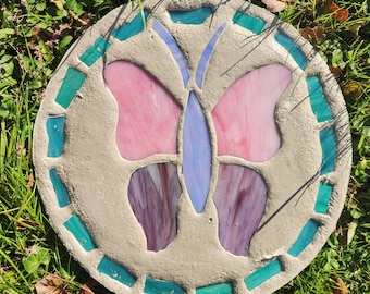 May include: A circular concrete stepping stone featuring a mosaic butterfly design. The butterfly has light pink upper wings, purple lower wings, and a light blue body. The border is decorated with rectangular teal glass tiles.