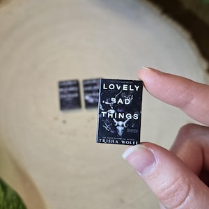 May include: Miniature book titled "LOVELY BAD THINGS" by Trisha Wolfe. The cover is black with white text and a graphic of a skull. Two other blurred books are in the background. The book is held by a person.