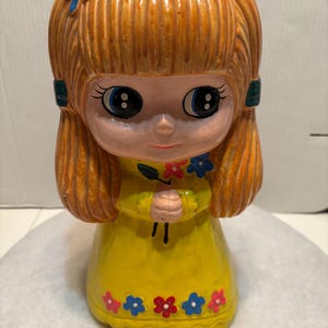 May include: A ceramic piggy bank shaped like a girl with long blonde hair, wearing a yellow dress with blue and red flowers. She is holding a flower in her hands and has big blue eyes.