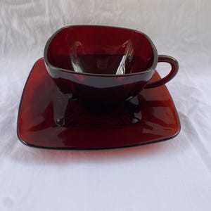 May include: A square red glass teacup and saucer set. The cup has a handle and the saucer is slightly larger than the cup.