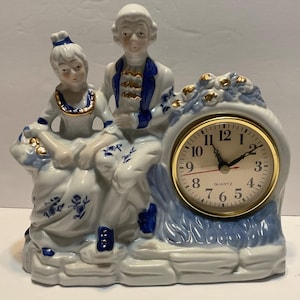 May include: A white porcelain clock featuring a seated couple in period attire. The clock face has gold trim and black numerals. The figures and clock base are decorated with blue floral patterns and accents.