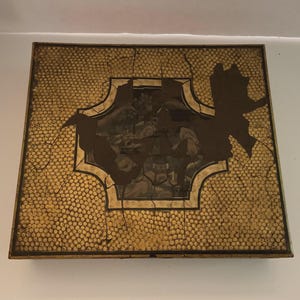 May include: Square, antique-style box with a gold, textured exterior and a central, ornate design. The box has a dark, decorative border and a cracked, aged appearance, suggesting it is vintage.